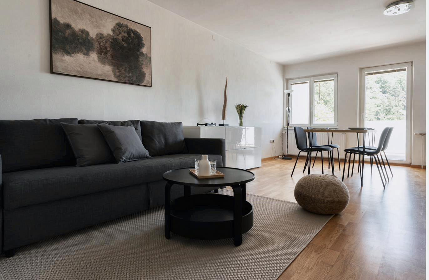 Wohnraum mit Sofa, Couchtisch und Essbereich vor hellen Fenstern